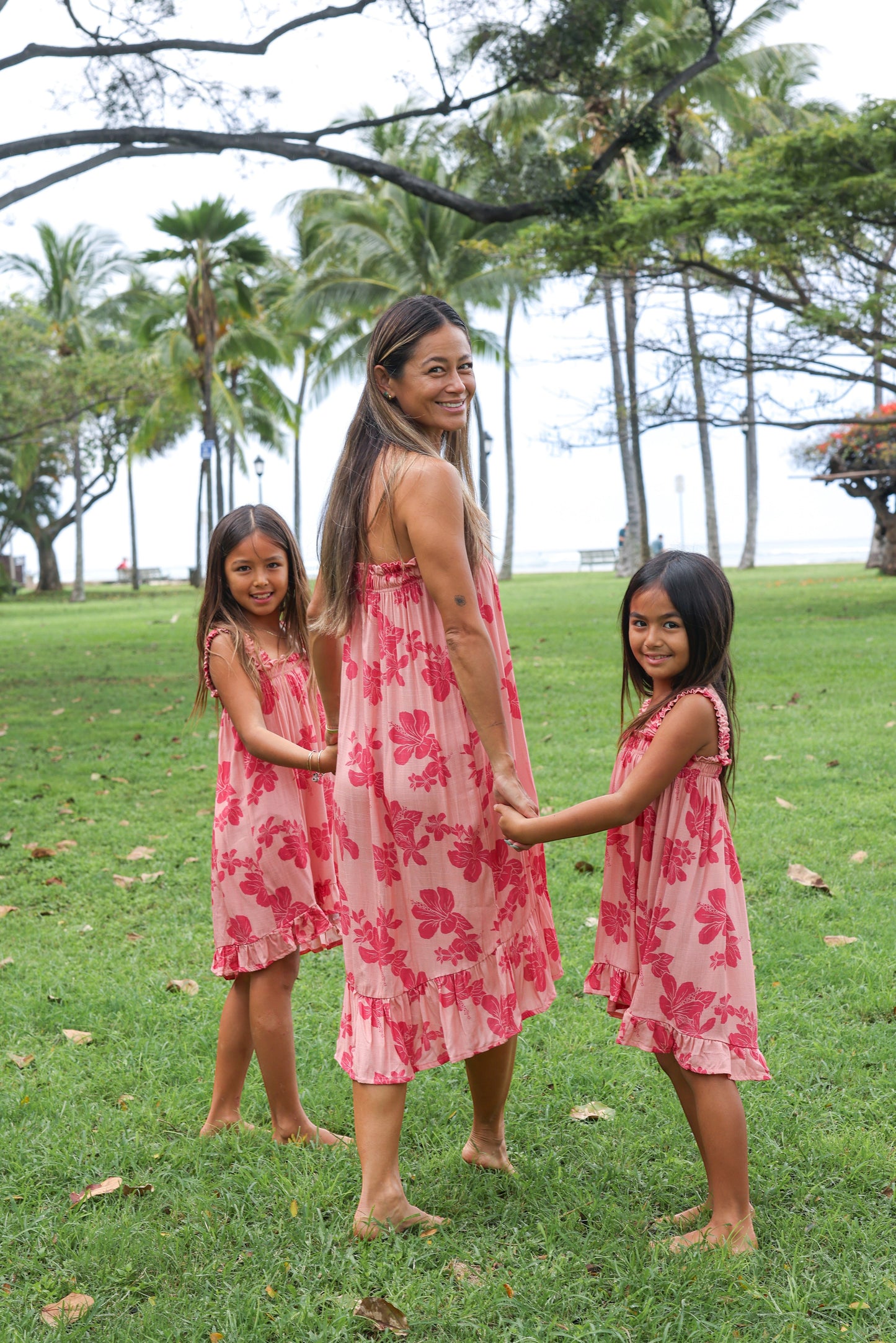 Guava Jammies Mahealani Mini Dress in coral pink with hibiscus print, featuring a ruffled hem and spaghetti straps. A mother and two daughters wear matching dresses.