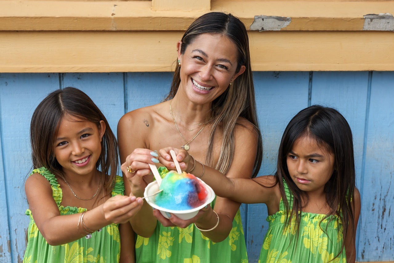 Guava Jammies Mahealani Mini Dress in green with yellow floral print, worn by a woman and two girls sharing rainbow shave ice.