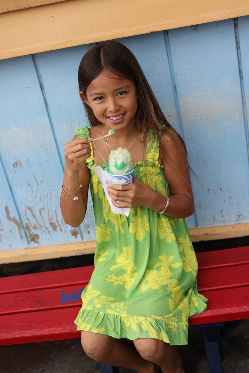Guava Jammies Mahealani Mini Dress in lime green with yellow floral print, featuring ruffled straps and hem. A young girl eats shaved ice.