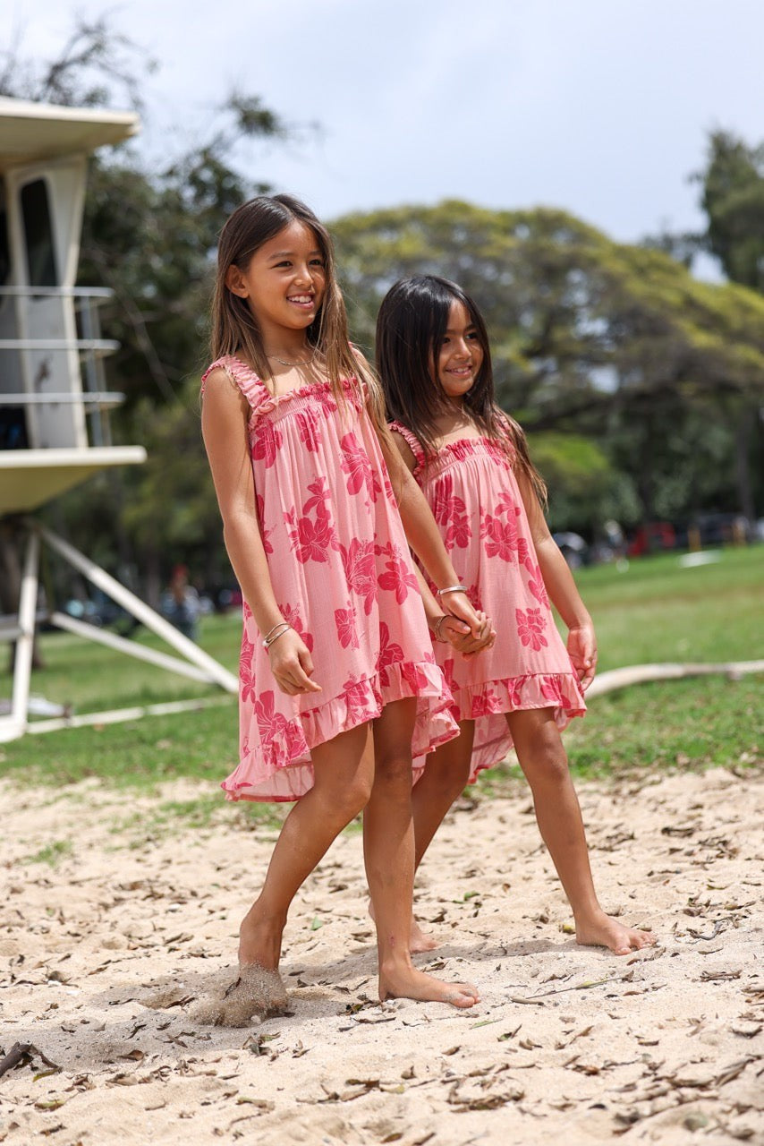 Guava Jammies Mahealani Mini Dress in pink with hibiscus print, featuring ruffled neckline and hem, on two girls at the beach.