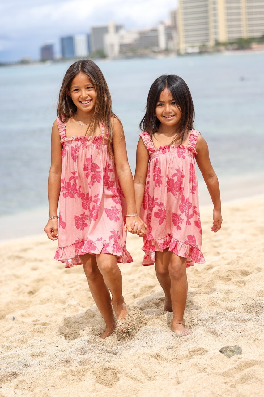 Guava Jammies Mahealani Mini Dress in pink with hibiscus print, featuring ruffled straps and hem. Two girls on a beach.