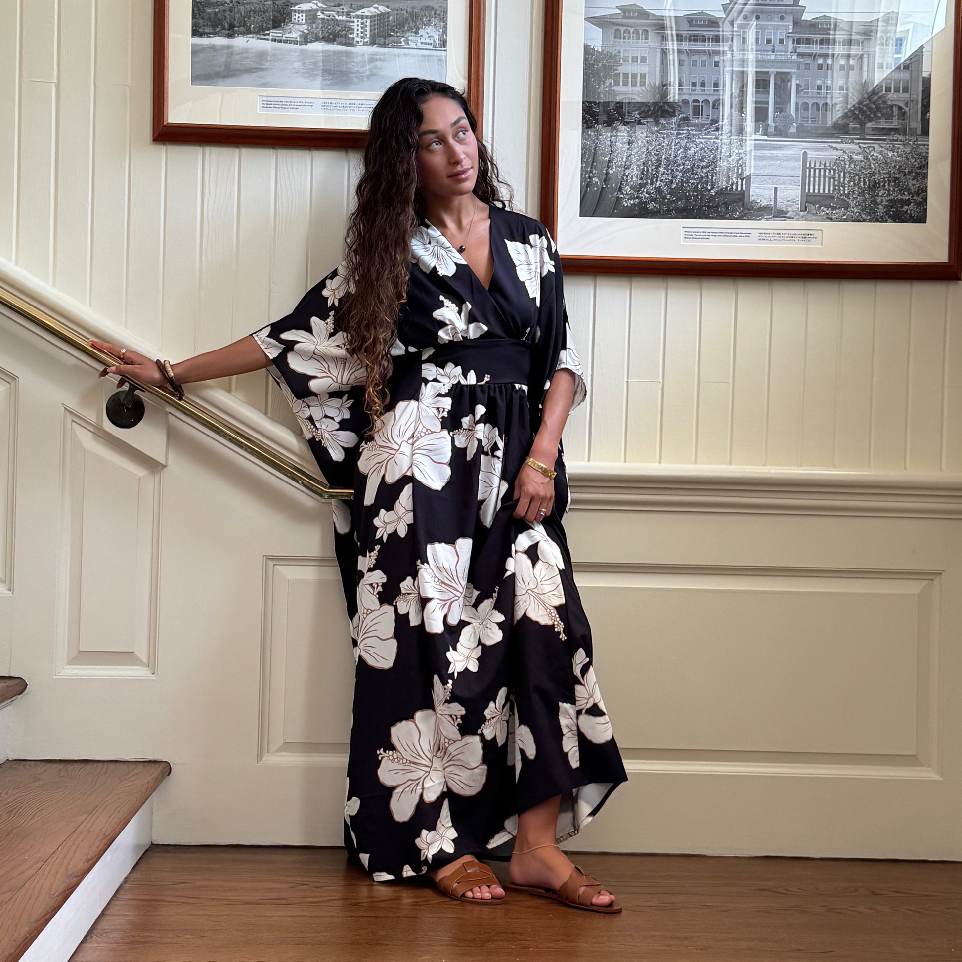 Guava & Co Poema Island Gown in black with white hibiscus print. Features a V-neck, cinched waist, and flowy skirt.